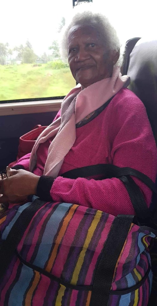An elderly woman seated on a bus, wearing a pink sweater and a scarf, with a colorful striped bag beside her.